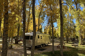 Camping trailer parked at site in the shade during fall surrounded by trees with hookups and picnic bench at Rio Chama RV Park in Chama, New Mexico.